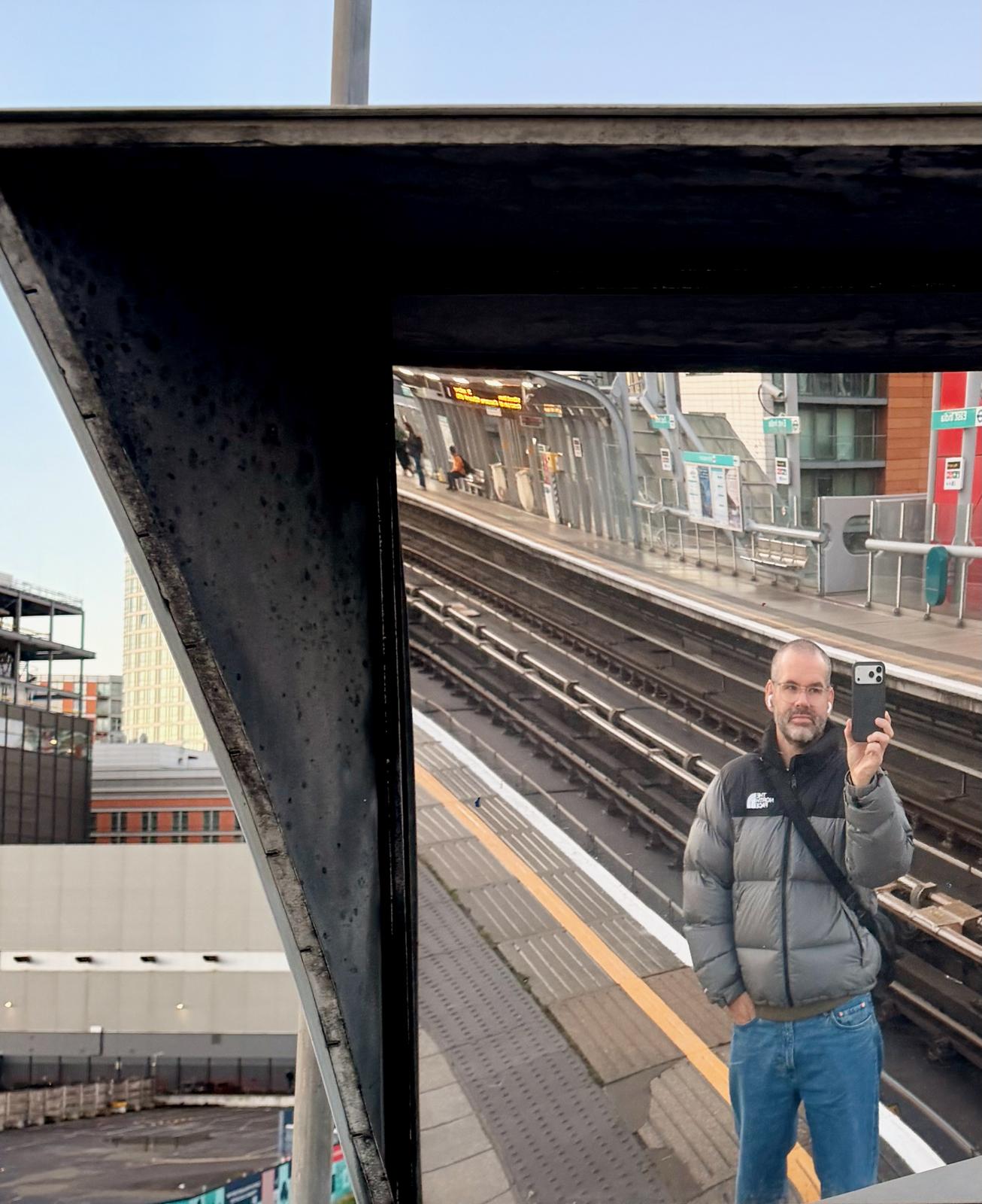 Me taking a mirror selfie in a mirror on a DLR platform while wearing a The North Face Nuptse puffer in grey
